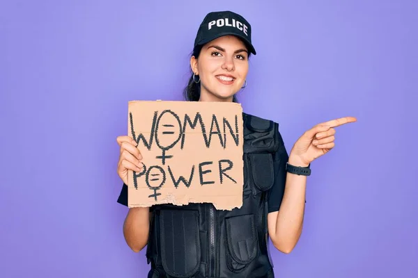 Police woman wearing security bulletproof vest uniform holding woman power protest cardboard very happy pointing with hand and finger to the side