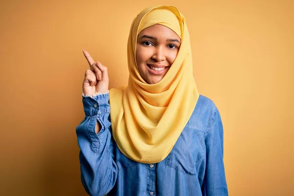 黄色の背景にアラブの伝統的なヒジャーブを身に着けている巻き毛を持つ若い美しい女性は 成功したアイデアで指を指しています 興奮し 第一番 — ストック写真