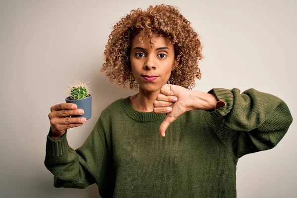 Young african american curly woman holding small cactus pot over isolated white background with angry face, negative sign showing dislike with thumbs down, rejection concept