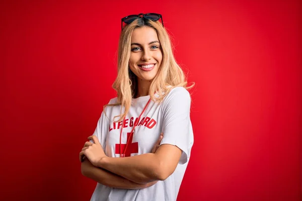 Young beautiful blonde lifeguard woman wearing t-shirt with red cross using whistle happy face smiling with crossed arms looking at the camera. Positive person.