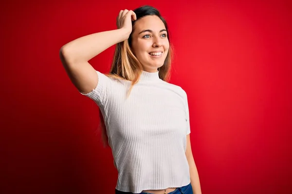 Beautiful blonde woman with blue eyes wearing casual white t-shirt over red background smiling confident touching hair with hand up gesture, posing attractive and fashionable
