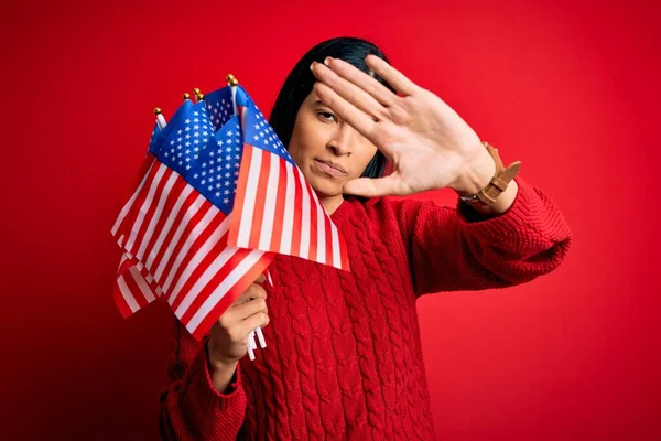 Young beautiful hispanic woman holding United States of American flag at independence day with open hand doing stop sign with serious and confident expression, defense gesture