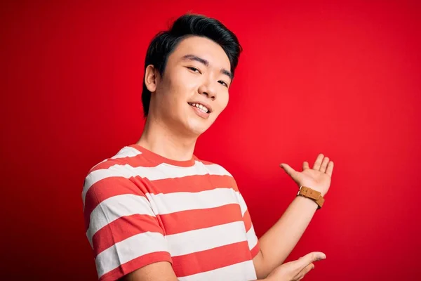 年轻英俊的中国男人穿着宽松的条纹T恤 站在红色的背景上 带着开放的手 自然地微笑着走进来 — 图库照片