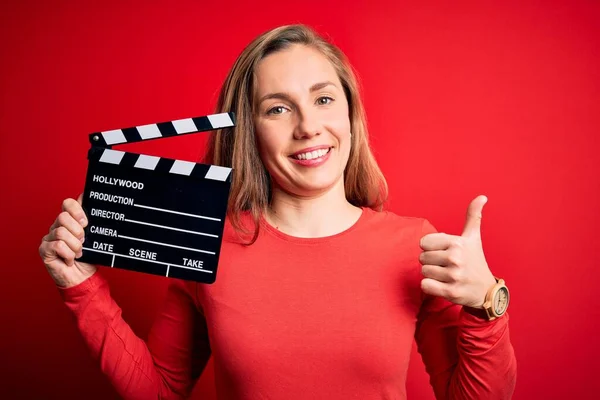 若いです美しいですブロンドClapper女性保持Clapboard上の隔離された赤の背景幸せなです大きな笑顔行いますOkサイン 親指アップとともに指 優れたサイン — ストック写真