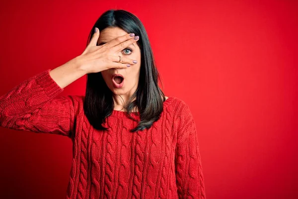 Young brunette woman with blue eyes wearing casual sweater over isolated red background peeking in shock covering face and eyes with hand, looking through fingers with embarrassed expression.
