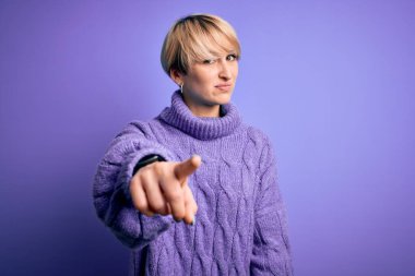 Kısa saçlı, balıkçı yaka kazak giyen, mor arka planda rahatsız edici ve hayal kırıklığına uğramış genç bir kadın. Sana kızgın ve öfkeli.