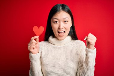 Genç Asyalı kadın kırmızı kalp kağıdını kırmızı arka planda tutarak gururla bağırıyor ve zaferi ve başarıyı kutluyor. Çok heyecanlı, neşeli bir duygu.