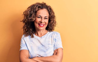 Orta yaşlı güzel bir kadın sarı arka planda durup kollarını kavuşturup kameraya bakarken gülümseyen günlük boyalı tişört giyiyor. Pozitif kişi.