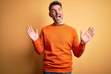 Turuncu kazak giyen yakışıklı genç adam izole edilmiş sarı arka plan üzerinde duruyor çılgın ve sinirli bir şekilde bağırıyor ve agresif bir ifade ve kollar havada bağırıyor. Hayal kırıklığı konsepti.