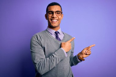 Yakışıklı Afro-Amerikan iş adamı gözlük takıyor ve mor arka planda kravat takıyor gülümsüyor ve iki eliyle ve parmaklarıyla kameraya bakıyor..