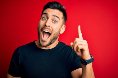 Siyah günlük tişörtler giyen genç yakışıklı adam izole edilmiş kırmızı arka planda dikiliyor ve başarılı bir fikri işaret ediyor. Heyecanlı ve mutlu. Bir numara..