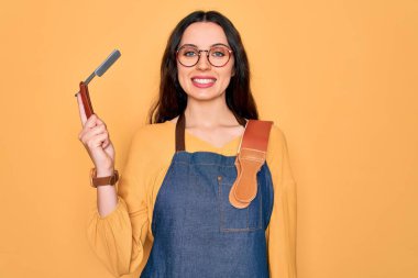Önlük takan genç, güzel bir berber kadın. Sarı arka planda jilet taşıyor. Mutlu bir yüzü var.