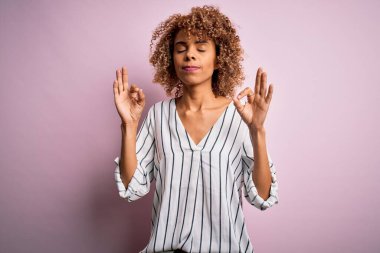 Güzel, kıvırcık saçlı, pembe arka planda çizgili tişört giyen, gözleri kapalı, meditasyon yaparken parmaklarıyla gülümseyen Afro-Amerikan kadını. Yoga konsepti.