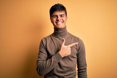 Genç yakışıklı bir adam, izole edilmiş sarı arka plan üzerine balıkçı yaka kazak giyiyor. Yüzünde mutlu ve doğal bir ifadeyle el ve parmak ile gülümseyen bir yüz ifadesi var.