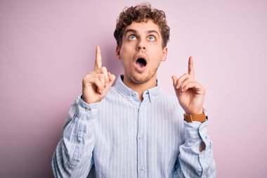 Sarışın, kıvırcık saçlı, beyaz arka planda çizgili gömlek giyen genç, yakışıklı adam şaşırmış ve şaşırmış bir şekilde yukarı bakıyor ve parmaklarıyla işaret ediyor ve kollarını kaldırıyor..