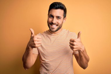 Sarı arkaplan işareti üzerine günlük çizgili tişört giyen sakallı yakışıklı genç adam el hareketleriyle olumlu jest yapıyor, gülümsüyor ve mutlu. Neşeli ifade ve kazanan jest.