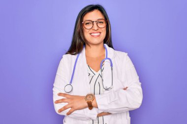 Mor arka planda steteskop ve tıbbi ceket giyen, kollarını kavuşturup kameraya gülümseyen profesyonel doktor kadın. Pozitif kişi.