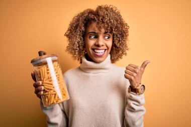 Genç Afro-Amerikan kıvırcık kadın elinde kavanozla İtalyan makarnası tutuyor ve gülümseyen bir gülümsemeyle başparmağıyla işaret ediyor.