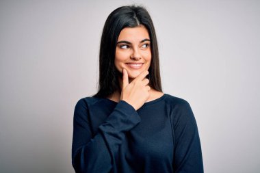 Genç, güzel esmer kadın beyaz arka planda dikilmiş, elleri çenesinde soru soran, düşünceli bir ifade takınıyor. Düşünceli bir yüzle gülümsüyor. Şüphe kavramı.