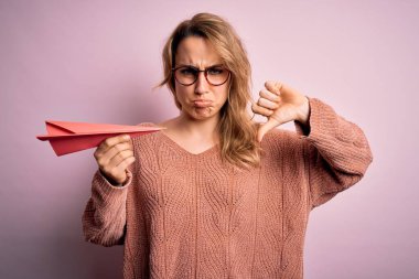 Genç, sarışın, gözlüklü, pembe arka planda kağıt uçak tutan, kızgın yüzlü, negatif işaretli, baş parmakları aşağıda, reddedilme konsepti olan güzel bir kadın.