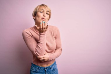 Kısa saçlı, balıkçı yaka kazak giyen, pembe arka planda kameraya bakıp havada öpücükler saçan, güzel ve seksi bir kadın. Aşk ifadesi.