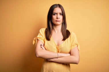 Sarı arka plan kuşkucu ve gergin, kollarını çapraz yapmış, güzel, esmer, sıradan bir elbise giymiş genç bir kadın. Negatif kişi.