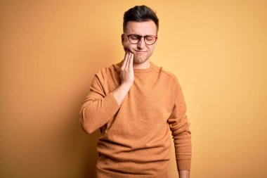 Gözlük takan yakışıklı beyaz bir adam ve sarı arka plan üzerinde sıradan bir kazak diş ağrısı ya da diş hastalığından dolayı acı dolu bir ifadeyle ağıza dokunuyor. Dişçi