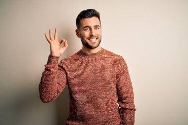 İzole edilmiş beyaz arka plan üzerinde duran sıradan bir süveter giyen genç yakışıklı olumlu gülümsüyor ve el ve parmaklarıyla 