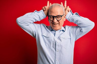 Orta yaşlı, yakışıklı, beyaz gömlekli ve gözlüklü, kırmızı arka planda komik ve çılgın pozlar veren, parmakları kafasında tavşan kulaklı, neşeli gülümseyen bir adam.