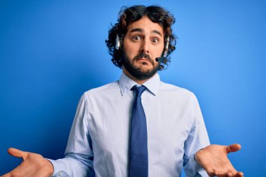 Sakallı genç, yakışıklı bir çağrı merkezi ajanı mavi arka planda kulaklık kullanıyor ve elleri ve kolları havada, kafası karışmış bir ifade takınıyor. Şüphe kavramı.