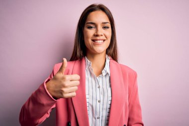 İzole edilmiş pembe arka planda duran ceketli güzel esmer kadın mutlu bir el hareketi yapıyor. Kameraya bakıp başarıyı gösteren ifade onaylanıyor.