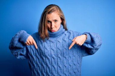 Mavi arka planda balıkçı yaka kazak giyen genç, güzel sarışın kadın üzgün ve üzgün görünüyor, parmaklarıyla yön gösteriyor, mutsuz ve depresif..