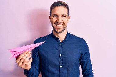 Pembe kağıt uçak tutan yakışıklı genç adam izole edilmiş arka planda duruyor ve pozitif ve mutlu görünüyor. Kendine güvenen bir gülümsemeyle dişlerini gösteriyor.