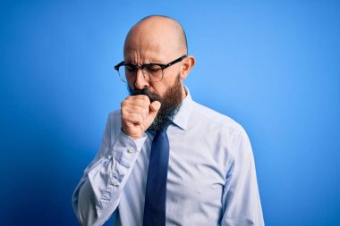 Sakallı, zarif kravat takan ve mavi arka planda gözlük takan yakışıklı kel bir adam kendini kötü hissediyor ve soğuk algınlığı veya bronşitin belirtisi olarak öksürüyor. Sağlık hizmeti kavramı.