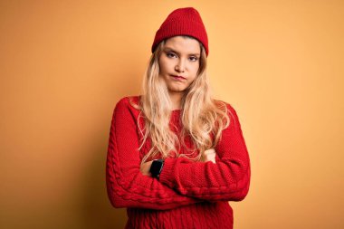 Genç, güzel, sarışın bir kadın, beyaz arka plan kuşkucu ve gergin, kolları çaprazlanmış bir yüz ifadesini tasvip etmeyen günlük kazak ve yün şapka giyiyor. Negatif kişi.