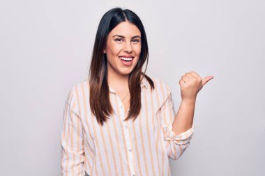 Genç, güzel, esmer, sıradan çizgili gömlek giyen, beyaz arka planda duran, baş parmağını yana doğru gösteren, ağzı açık gülümseyen bir kadın.