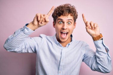 Sarışın, kıvırcık saçlı, beyaz arka planda çizgili tişört giyen, şaşkın, şaşkın, parmaklarıyla işaret eden ve kollarını kaldıran genç bir adam..