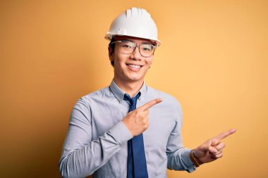 Güvenlik kaskı takan ve sarı arka planda kravat takan genç yakışıklı mimar gülümseyip kameraya bakıyor ve iki eliyle ve parmaklarıyla kenarı işaret ediyor..