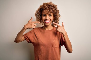 Güzel Afro-Amerikalı kadın kıvırcık saçlı beyaz arka plan üzerine günlük tişörtler giyip gülümseyen ve parmakları, dişleri ve ağzı ile işaret eden. Diş sağlığı konsepti.