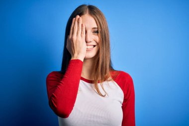 Genç, güzel, kızıl saçlı bir kadın, izole edilmiş mavi arka plan üzerine günlük tişört giyiyor. Bir gözünü eliyle kapatıyor. Yüzünde kendine güvenen bir gülümseme ve sürpriz bir duygu var..