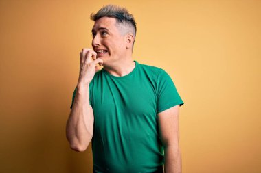 Sarı arka plan üzerine gündelik yeşil tişört giyen genç ve modern adam elleri ağız yiyen tırnaklarla gergin ve stresli görünüyor. Endişe sorunu.