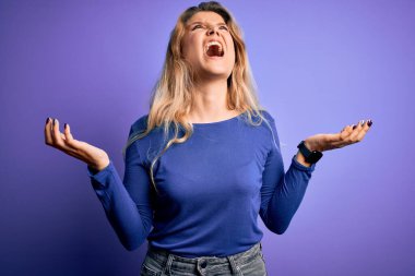Genç, güzel, sarışın bir kadın, izole edilmiş mor arka plan üzerine gündelik tişört giyip deli gibi bağırıyor ve agresif bir ifade ve kollarla bağırıyor. Hayal kırıklığı konsepti.