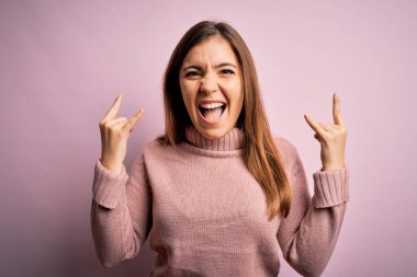 Güzel genç bir kadın pembe izole edilmiş arka plan üzerine balıkçı yaka kazak giyip çılgın bir ifadeyle elleri havada rock sembolü yaparak bağırıyor. Müzik yıldızı. Ağır konsept.