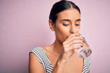 Genç, güzel esmer kadın, ferahlamak için bir bardak sağlıklı su içiyor. İzole edilmiş pembe arka planda duruyor.