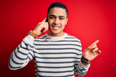 Brezilyalı bir adam akıllı telefondan konuşuyor izole edilmiş kırmızı arka planda çok mutlu bir şekilde el ve parmak kenarı işaret ediyor.