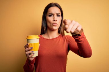 Genç ve güzel esmer kadın sarı arka planda kahve içerek kamerayı işaret ediyor ve sen, el işareti, ön taraftan pozitif ve kendinden emin bir hareket.
