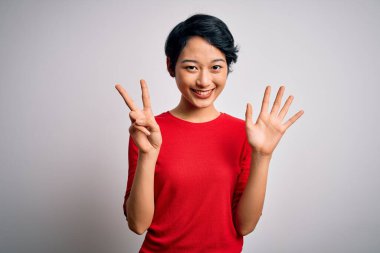 Genç, güzel Asyalı bir kız, günlük kırmızı tişört giyiyor. İzole edilmiş beyaz arka plan üzerinde duruyor ve yedinci parmaklarıyla işaret ediyor. Kendinden emin ve mutlu bir şekilde gülümsüyor..