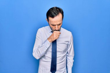 Soyutlanmış mavi arka plan üzerinde duran şık kravat takan orta yaşlı yakışıklı iş adamı kendini iyi hissetmiyor ve soğuk algınlığı veya bronşitin belirtisi olarak öksürüyor. Sağlık hizmeti kavramı.