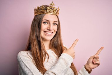 Genç, güzel, kızıl saçlı, Kraliçe tacını takmış pembe arka planda gülümseyen ve kameraya bakan iki eliyle ve parmaklarıyla yüzünü gösteren bir kadın..