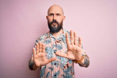 Sakallı ve dövmeli yakışıklı, pembe arka planda sıradan bir çiçek gömleği giyen, elleri hareket eden, reddeden ve iğrenç bir ifadeyle reddeden, kel bir adam. Dur ve yasakla.